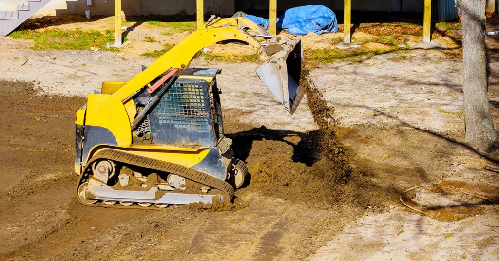 Yellow skid steer loader spreading soil to level ground during residential grading and landscaping work.